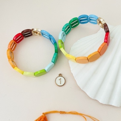 Dois braceletes coloridos ao lado de um medalhão branco com o número 1, sobre uma superfície clara com uma concha branca