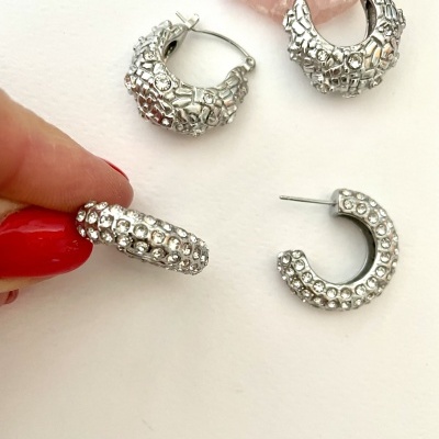 Quatro pares de brincos prateados com cristais, sobre fundo branco, uma mão segurando um brinco.