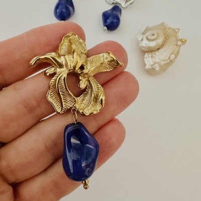 Brincos florais metálicos dourados e prateados com pendente azul, em mão sobre fundo branco.