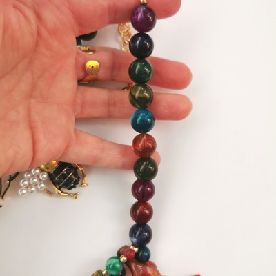 Colar multicolorido com contas redondas e pendentes em forma de galhos, segurado por mão em fundo branco