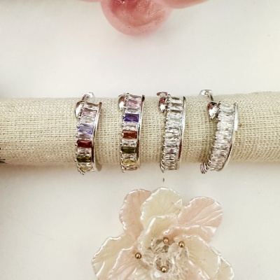 Quatro brincos prateados com pedras retangulares coloridas e transparentes em suporte bege com flor decorativa ao fundo.