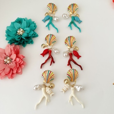 Três pares de brincos coloridos em forma de concha com peixes e ramos, ao lado de duas flores em tecido azul e rosa.