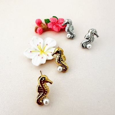 Brincos cavalo-marinho dourados e prateados com pérolas, sobre superfície clara com flores artificiais