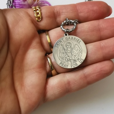Medalha redonda prateada de São Gabriel com texto, na mão com joias