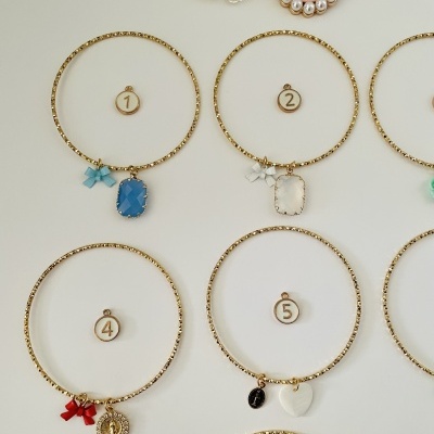 Braceletes dourados com pendentes variados e laços coloridos sobre fundo branco com broches decorativos