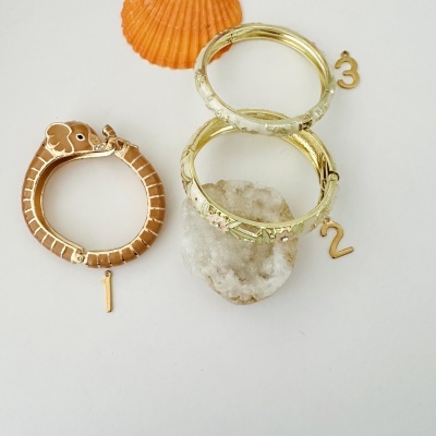 Pulseiras douradas decoradas, uma em forma de elefante, com conchas e pedra branca de fundo