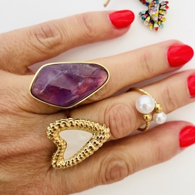 Mão com três anéis dourados com pedras e unhas vermelhas, broche colorido no fundo branco