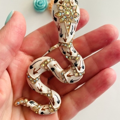 Broche de serpente branca com detalhes dourados e strass numa mão com unhas azuis claras