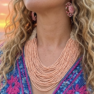 Colar de contas rosa claro com fecho de croché bege em mulher com cabelo loiro ondulado e vestido azul bordado
