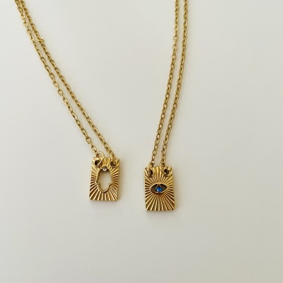 Dois colares dourados com pendentes retangulares e padrão de raios, um com espaço oval vazio e outro com olho azul.