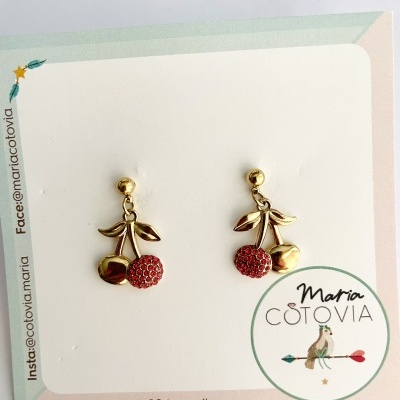 Brincos dourados em forma de cerejas com detalhes vermelhos em cartão branco com design verde e rosa