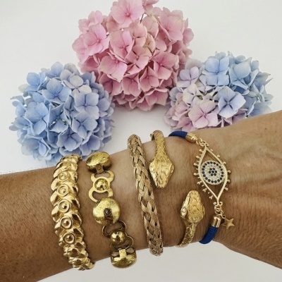 Braceletes dourados e azul com pingente olho grego num braço, flores hortênsia ao fundo