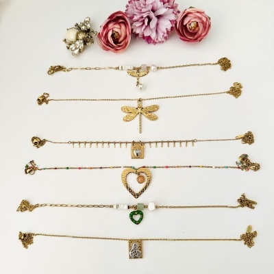 Sete pulseiras douradas com pendentes variados, sobre fundo branco com flores e broche ao fundo.