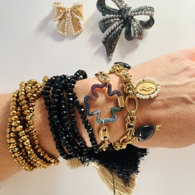 Pulseiras de contas douradas e pretas, bracelete com pendente colorido e pulseira dourada com pingente oval, com dois broches em forma de laço cinza e dourado com pérolas sobre fundo branco.