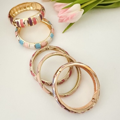 Conjunto de pulseiras douradas com detalhes coloridos, colocadas perto de flores cor-de-rosa