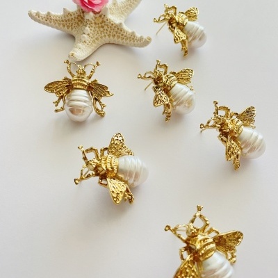 Brincos em forma de abelha com pérolas e metal dourado sobre fundo branco com estrela-do-mar e flor rosa