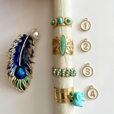 Anéis dourados com pedras turquesa num suporte de tecido bege, broche forma pena azul e medalhões numerados 1 a 4