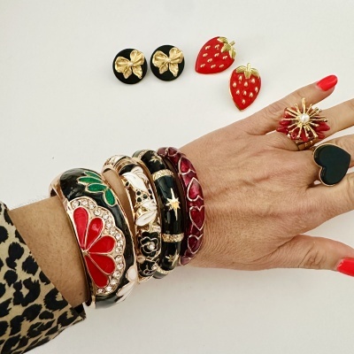 Pulseiras e anéis coloridos em esmalte com detalhes dourados numa mão, brincos de morango e flor ao fundo