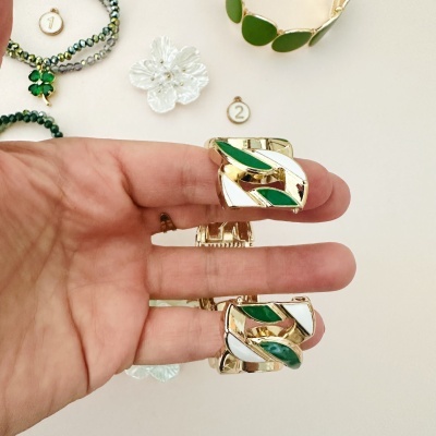 Brincos verdes e dourados com esmalte branco segurados numa mão sobre fundo claro com bijuterias numeradas.