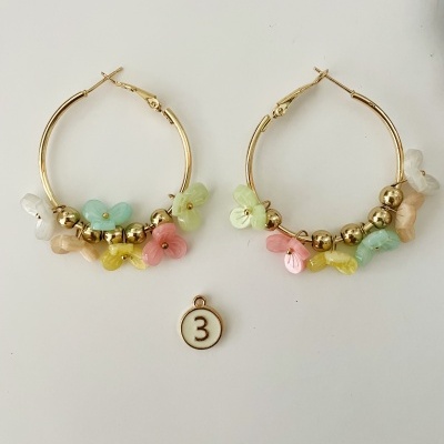 Brincos de argola dourados com flores coloridas e esferas douradas numa superfície branca