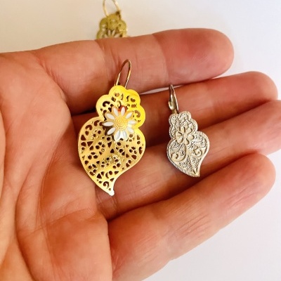 Par de brincos em forma de folhas, um dourado com flor branca e outro prateado gravado.
