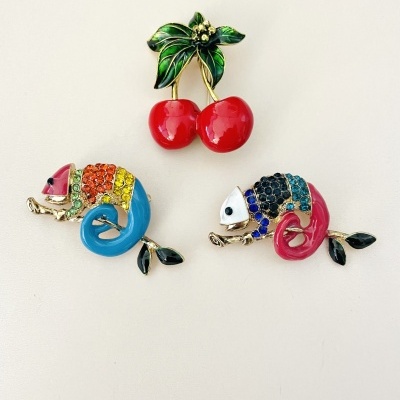 Três broches coloridos: dois camaleões brilhantes e um par de cerejas vermelhas com folhas verdes.