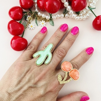 Mão com dois anéis grandes em cenário com colar de pérolas e cerejas decorativas