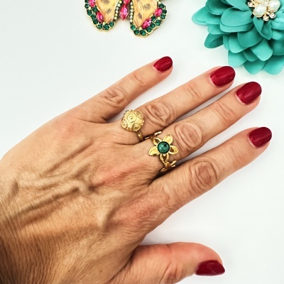 Mão com anéis dourados e unhas vermelhas junto a flor azul artificial e brinco de borboleta com pedras.
