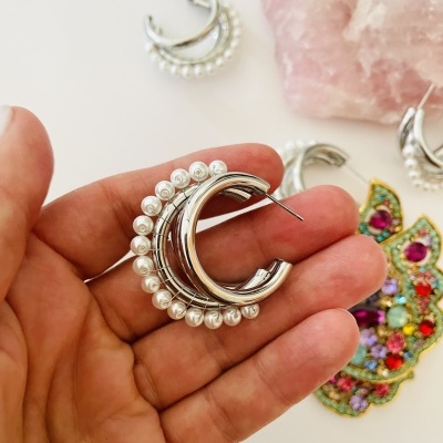Brincos de argola prateados com pérolas numa mão e elementos decorativos ao fundo
