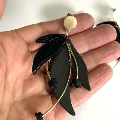 Brinco preto com detalhes dourados em forma de folha e flor, segurado numa mão.