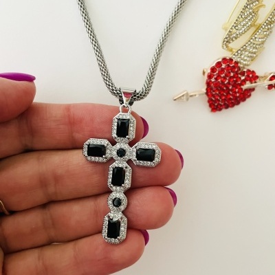 Colar com pendente em forma de cruz com pedras pretas e cristais, em fundo branco com clips dourados e coração vermelho