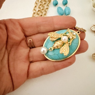Colar com pendente de pedra turquesa azul e abelha dourada com pérolas em fundo branco.
