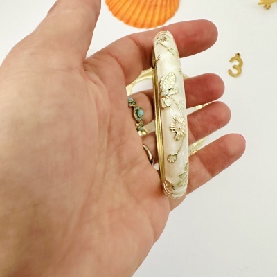Bracelete creme decorado com flores douradas segurado por mão com anel turquesa