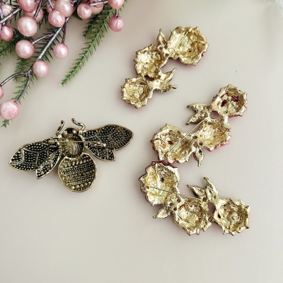 Broches dourados em forma de abelha e flores vistos de trás junto a ramos com bagas cor de rosa