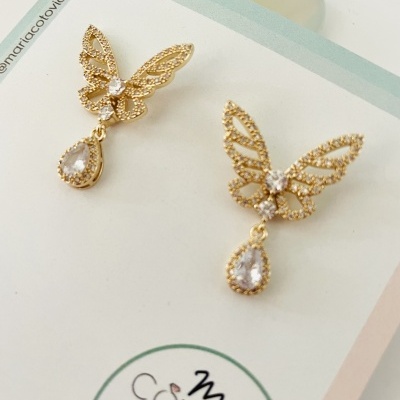 Brincos dourados de borboleta com pedras transparentes apresentados em cartão branco com texto
