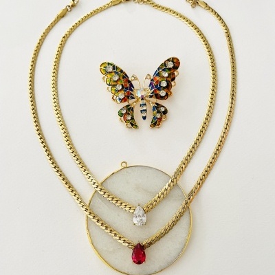 Dois colares dourados e um broche de borboleta colorida sobre fundo branco.