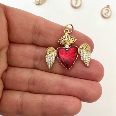 Pendente dourado com coração vermelho e asas segurado por mão