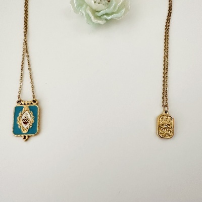 Dois colares dourados com pendentes retangulares, um com esmaltação azul e detalhes, outro com relevo de olho e coroa, sobre fundo branco.