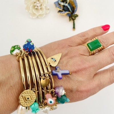 Mão com pulseiras douradas coloridas e anel com pedra verde sobre fundo branco com dois alfinetes de flor