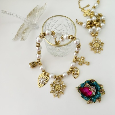 Colar de contas peroladas e pendentes dourados, vidro transparente com borda dourada, joia colorida e libélula translúcida sobre fundo branco.
