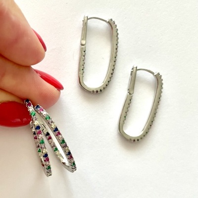 Par de brincos argola prateada com pedras coloridas, um deles segurado por mão com unhas vermelhas