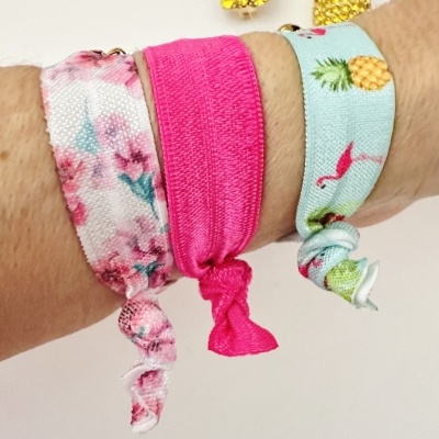 Três pulseiras coloridas de tecido amarradas no pulso, com padrão floral, rosa sólido e abacaxis com flamingos.