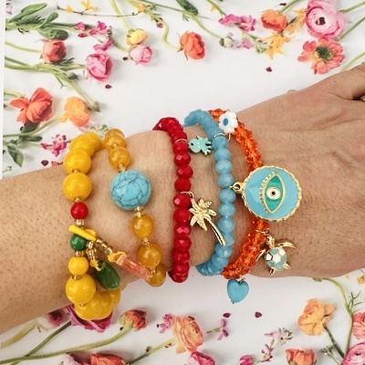 Pulseiras coloridas com pendentes diversas num braço sobre fundo de flores coloridas.