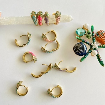 Brincos argola dourados com padrões coloridos em escama e um broche decorativo com conchas e pérolas