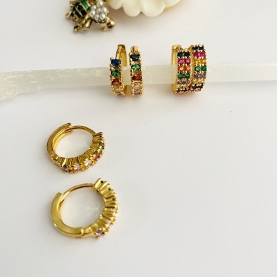 Quatro pares de brincos de argola dourados com pedras coloridas sobre fundo branco com flor decorativa.