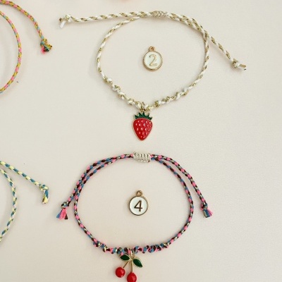 Pulseiras trançadas com pendentes de morango e cerejas, etiquetas numeradas 2 e 4, fundo claro com decoração