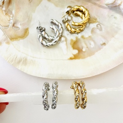 Brincos argola prateados e dourados com textura retorcida sobre concha e suporte branco