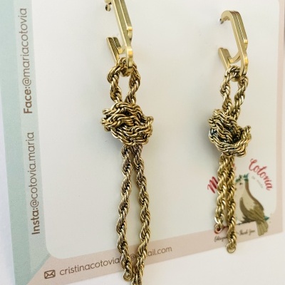 Brincos dourados com correntes torcidas e nó central em cartão de apresentação.