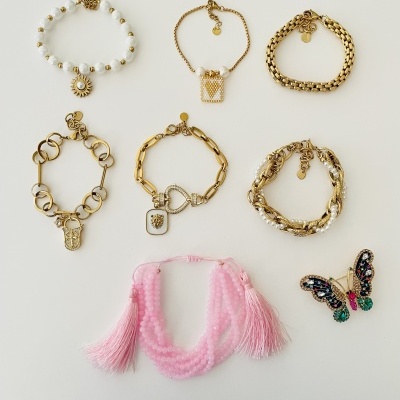 Sete acessórios de moda, incluindo pulseiras douradas e rosa e um alfinete de borboleta colorido, sobre fundo branco.