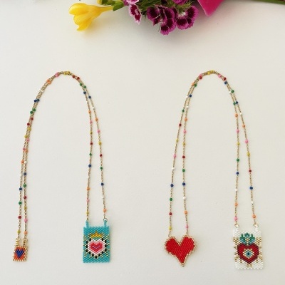 Quatro colares com pingentes de contas coloridas em forma de coração e quadrados, sobre fundo branco com flores.